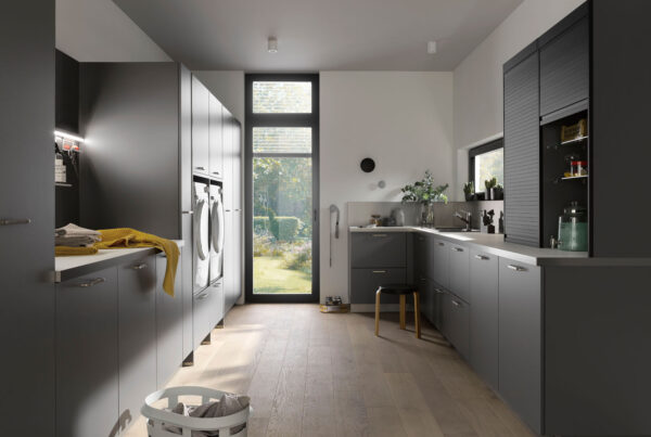 Moderne graue Waschküche mit Waschmaschinen, Trockner, großen Fenstern und Holzboden. Perfekt organisiert und gut belichtet mit eleganten Schränken.