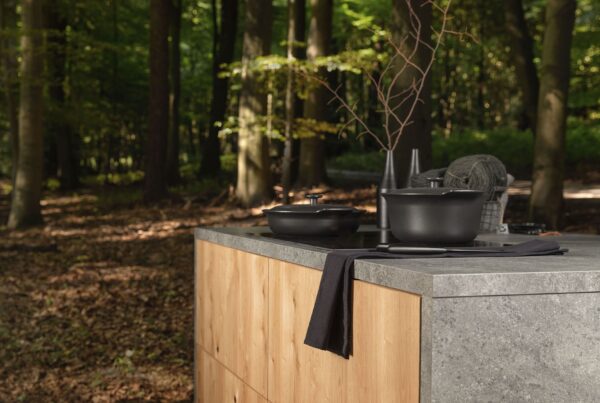 Kochinsel im Freien im Wald, mit Holz- und Steinoberfläche, schwarzem Topf und Tuch, umgeben von Bäumen und natürlichem Licht.
