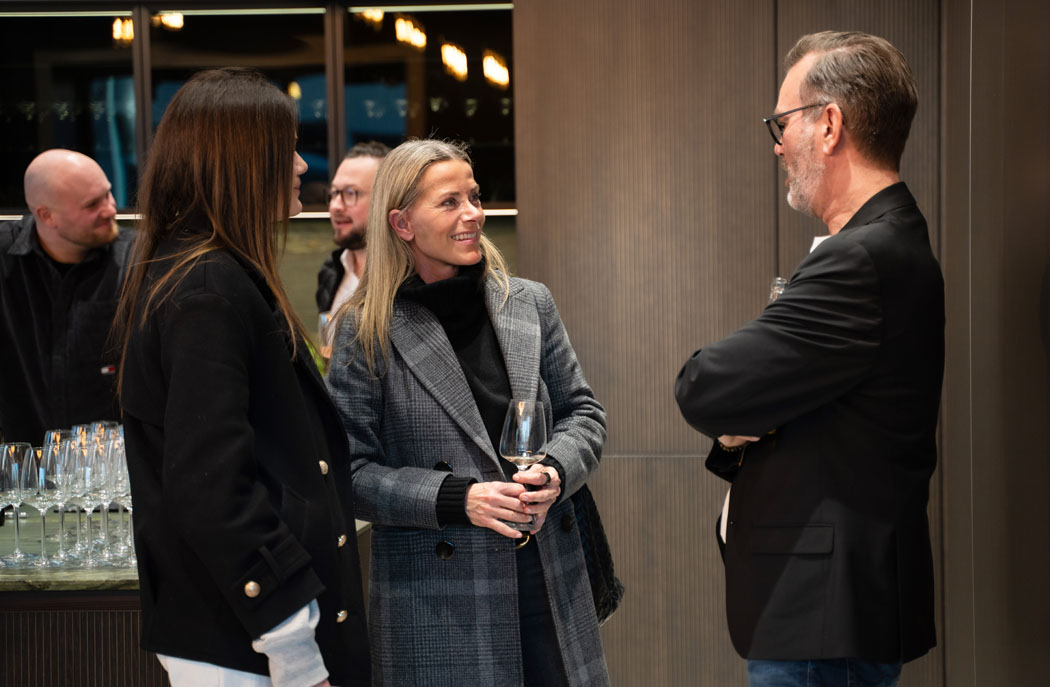 Drei Personen unterhalten sich in einem eleganten Raum mit Gläsern im Hintergrund, die eine freundliche Atmosphäre schaffen.