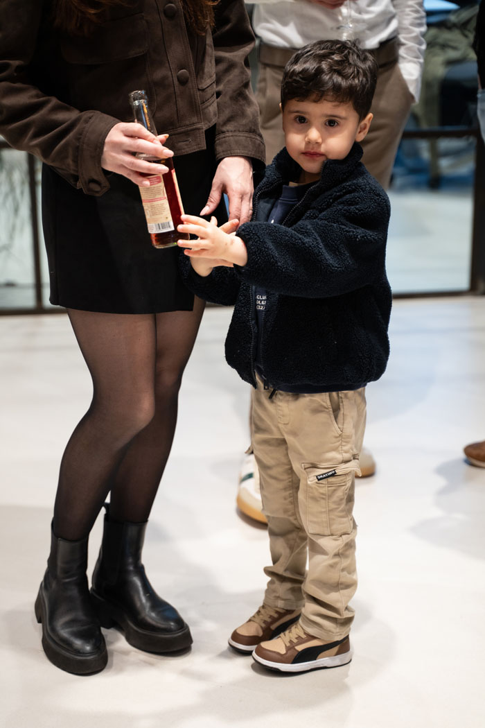 Ein kleiner Junge steht neben einer Frau und hält eine Flasche. Er trägt einen schwarzen Fleece und eine beige Hose.