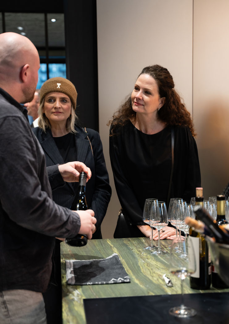 Eine Gruppe von Menschen diskutiert an einem Tisch mit Weingläsern und einer Flasche Wein in einem modernen Raum.