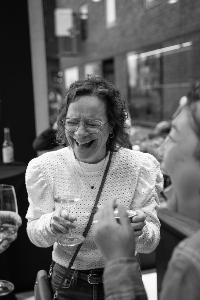 Eine lachende Frau mit Brille hält ein Weinglas und genießt einen fröhlichen Moment mit Freunden.