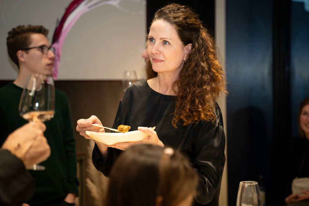 Eine Frau mit lockigem Haar hält einen Teller mit Essen während einer geselligen Veranstaltung, umgeben von Gästen.