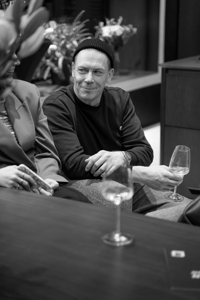 Ein Mann in einem schwarzen Pullover sitzt entspannt mit einem Glas Wein in der Hand, umgeben von Freunden.