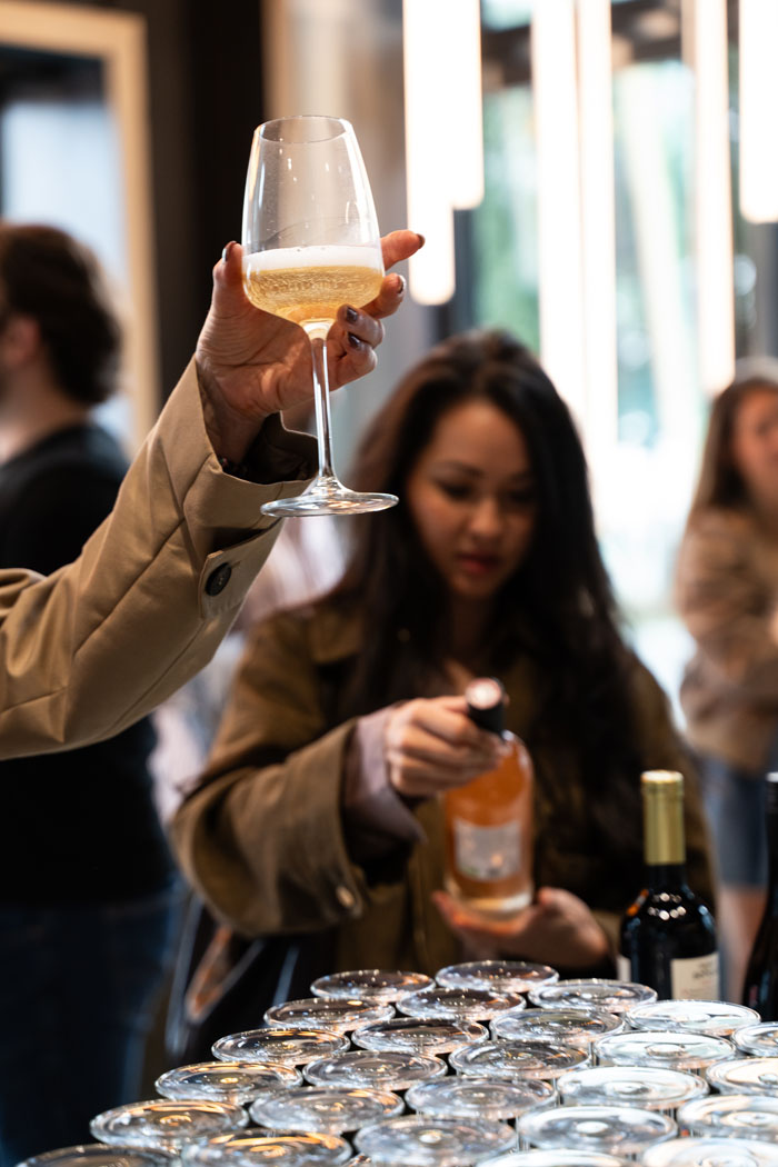 Eine Hand hält ein Glas Sekt hoch, während im Hintergrund eine Frau eine Flasche Wein öffnet und viele leere Weingläser auf einem Tisch stehen.