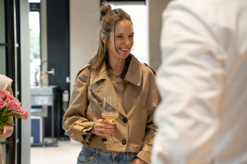 Eine lächelnde Frau mit hochgesteckten Haaren trägt einen beigen Trenchcoat und hält ein Weinglas in der Hand, während sie sich mit jemandem unterhält.