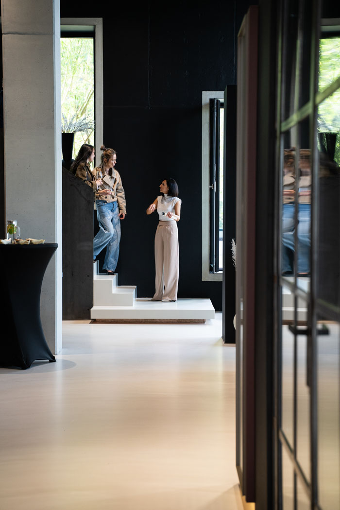 Drei Frauen unterhalten sich in einem modernen Raum mit schwarzer Wand und weißer Treppe, während im Vordergrund ein Stehtisch zu sehen ist.