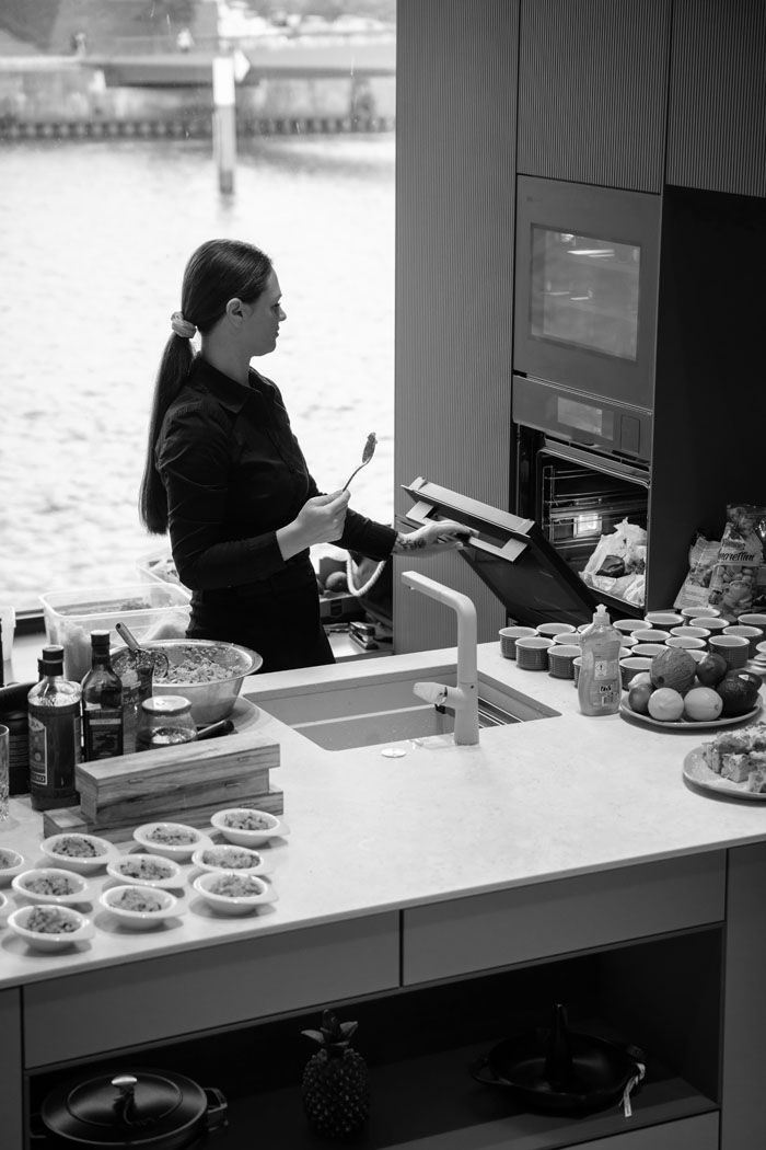 Eine Frau in schwarzer Kleidung steht in einer modernen Küche, hält einen Löffel und öffnet den Backofen, während viele kleine Schalen bereitstehen.