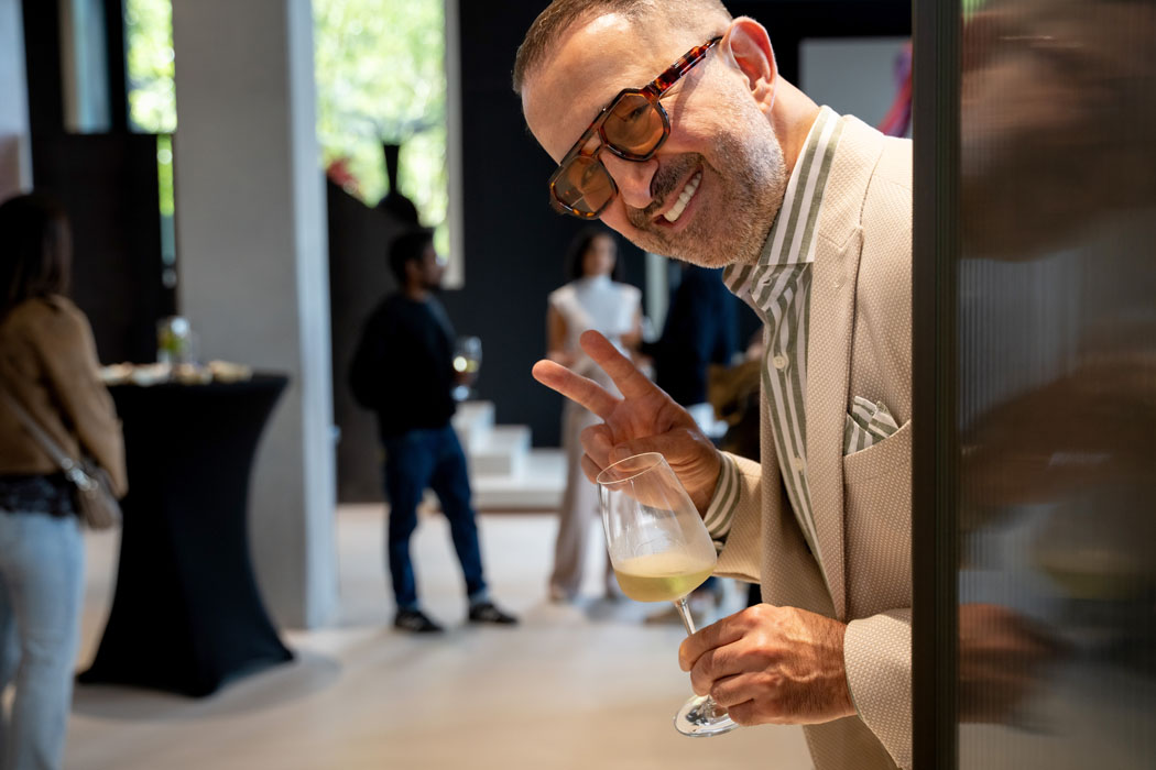 Ein lächelnder Mann mit Brille und beigem Sakko macht ein Friedenszeichen, während er ein Weinglas hält, im Hintergrund sind Gäste bei einer Feier.