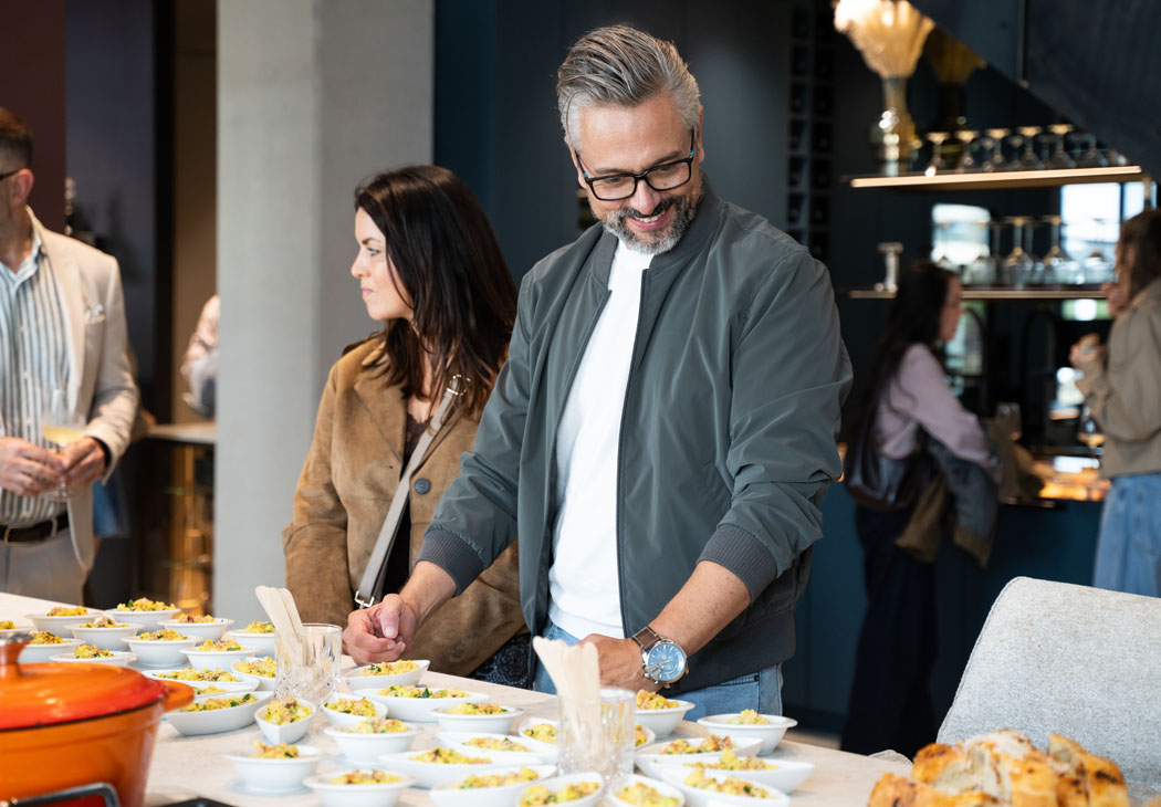 Ein lächelnder Mann mit Brille und grauer Jacke wählt bei einem geselligen Event kleine Häppchen von einem reich gedeckten Buffet aus.