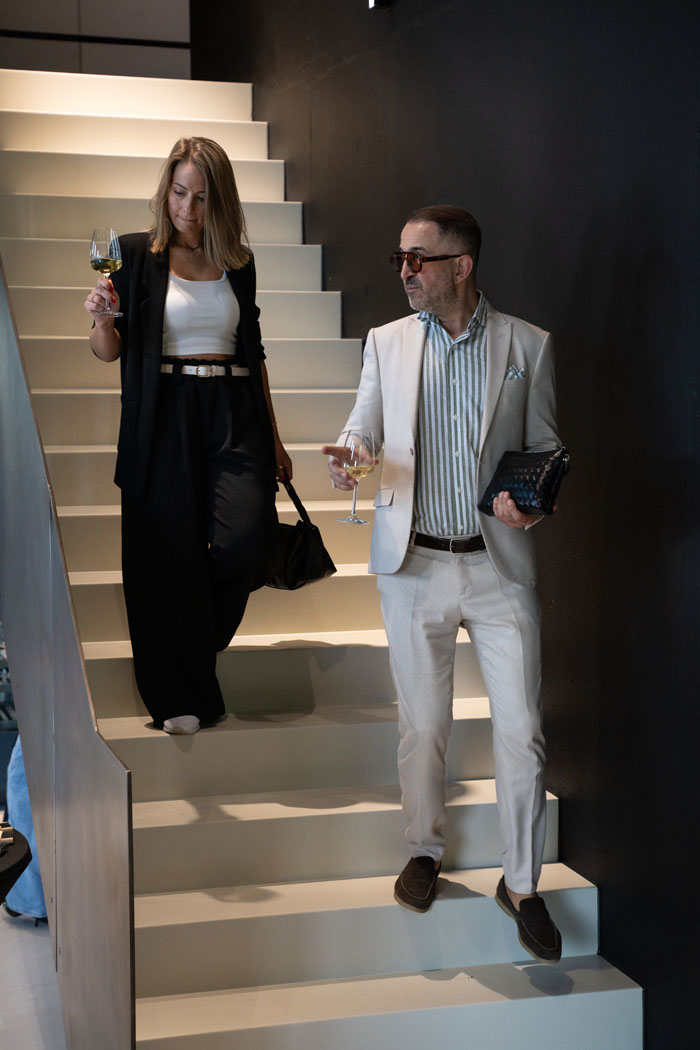 Eine Frau in schwarzem Outfit und ein Mann im hellen Anzug gehen eine Treppe hinunter, beide halten jeweils ein Glas Weißwein in der Hand.