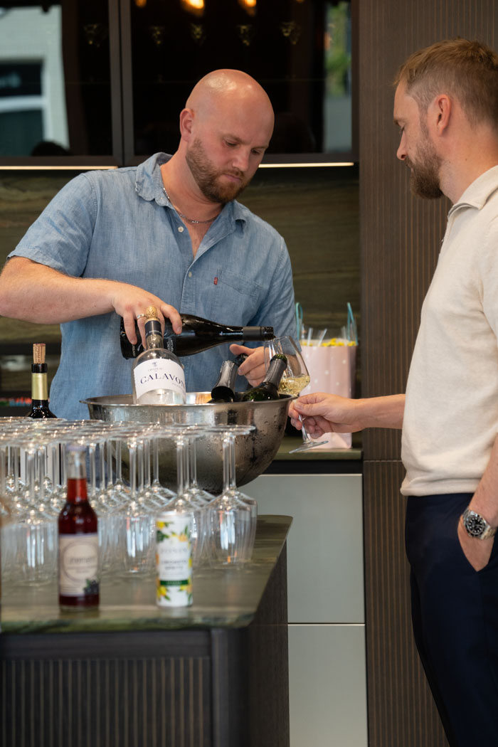 Ein Mann schenkt einem anderen in einer modernen Bar Weißwein aus einer Flasche Chateau Calavon in ein Glas ein, während viele Weingläser bereitstehen.