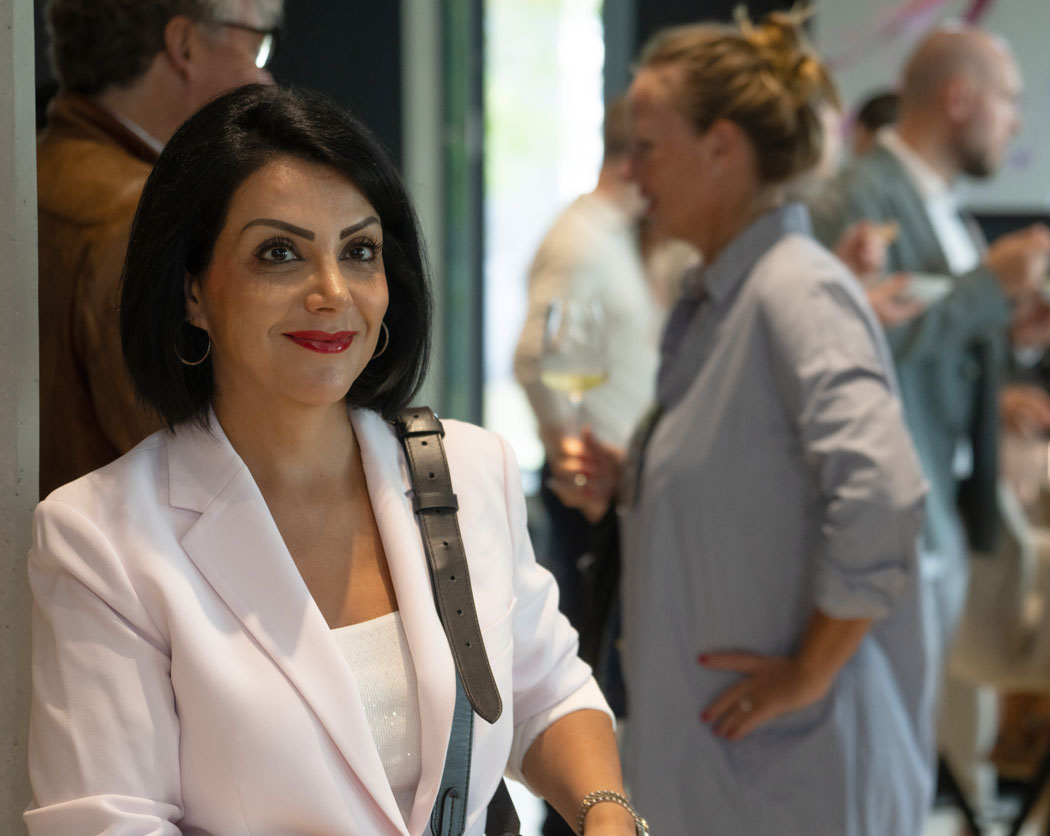 Eine elegant gekleidete Frau mit dunklem Haar und rotem Lippenstift lächelt in die Kamera, während im Hintergrund Gäste auf einer Feier stehen.
