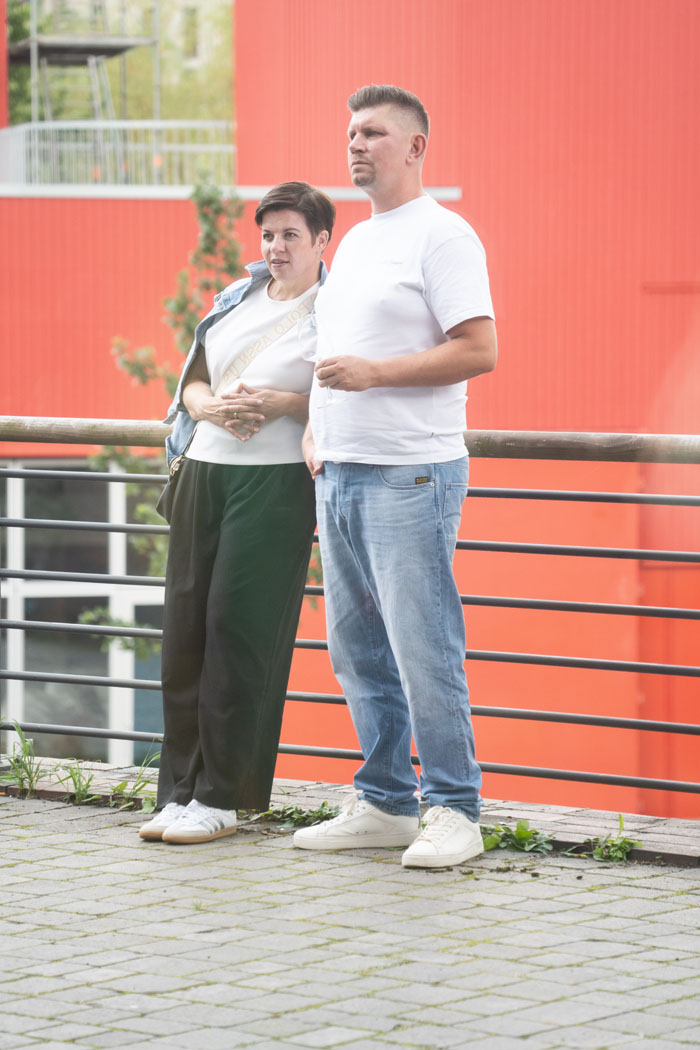 Ein Paar steht entspannt an einem Geländer vor einer leuchtend roten Wand. Beide tragen weiße T-Shirts und helle Sneaker in einer urbanen Umgebung.