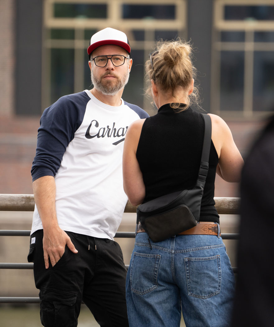 Ein bärtiger Mann mit Brille und Cap unterhält sich mit einer Frau, die ein schwarzes Top, eine Umhängetasche und eine blaue Jeans trägt, im Freien.