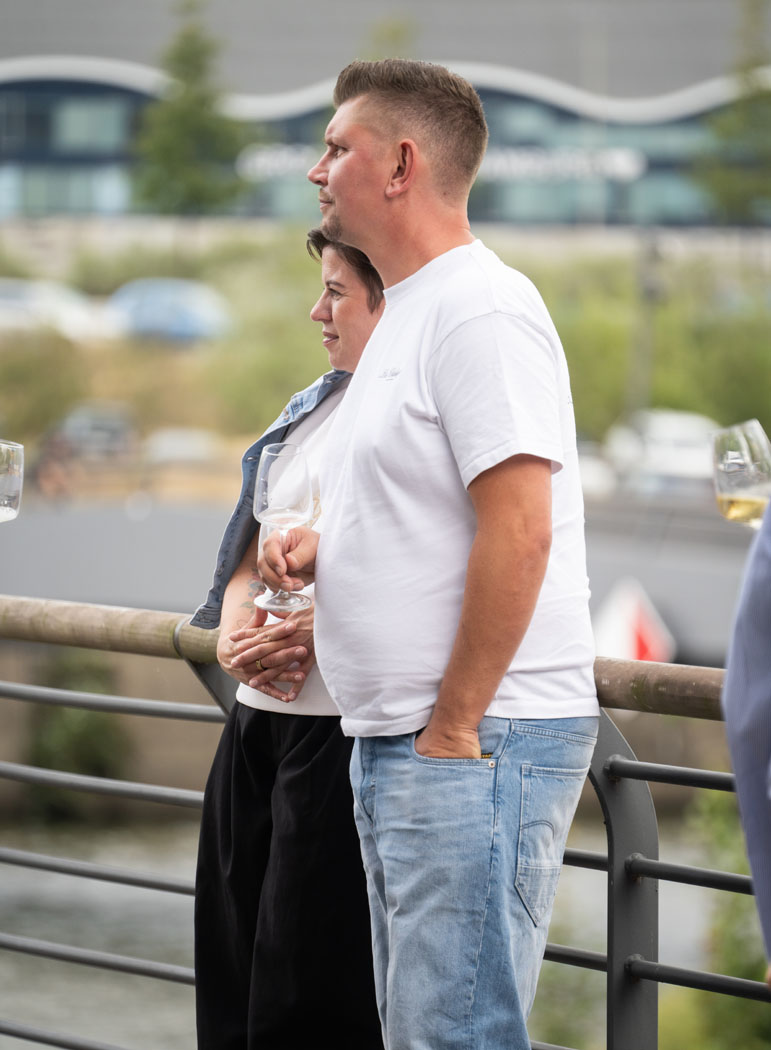Ein Paar steht an einem Geländer, hält Weingläser in der Hand und blickt gemeinsam in die Ferne, während sie entspannt Zeit im Freien verbringen.
