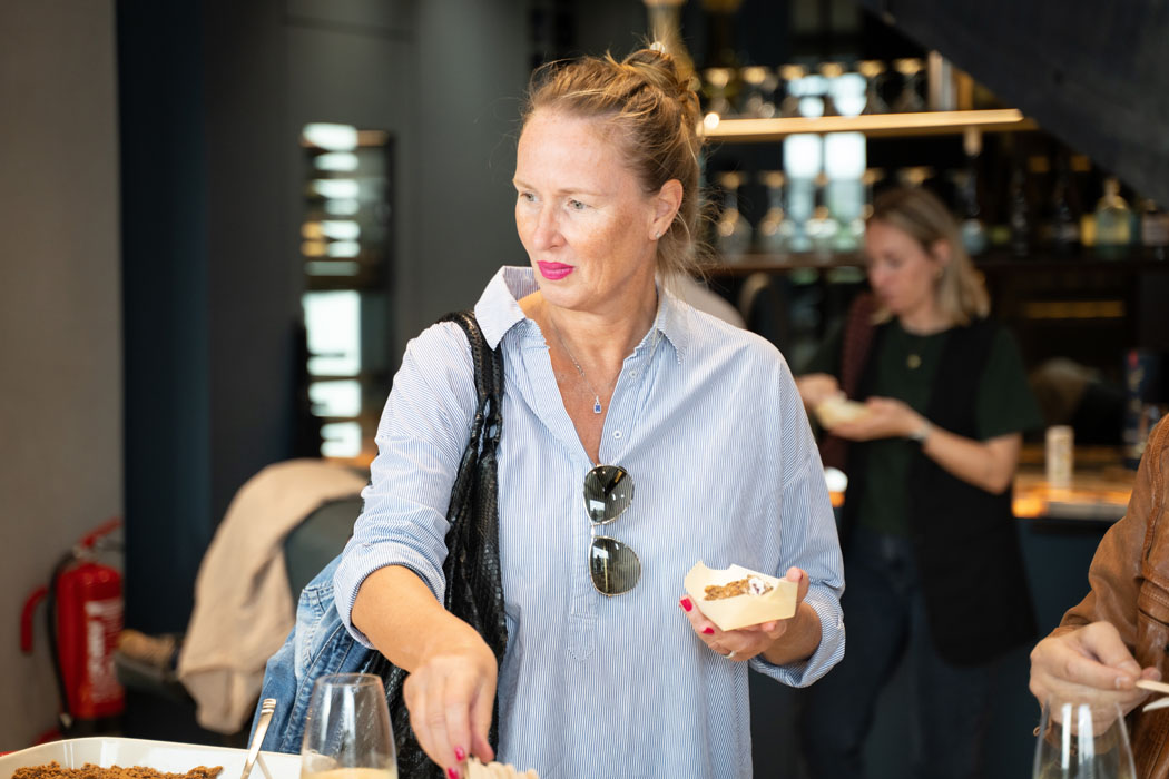Eine blonde Frau in einem hellblauen Hemd greift bei einem Buffet zu einem kleinen Snack, während im Hintergrund eine weitere Person zu sehen ist.