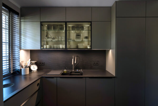 Moderne, dunkelgraue Küche mit beleuchteten Glasschränken, in denen weißes Geschirr steht, sowie einer dunklen Arbeitsplatte mit integriertem Spülbecken.