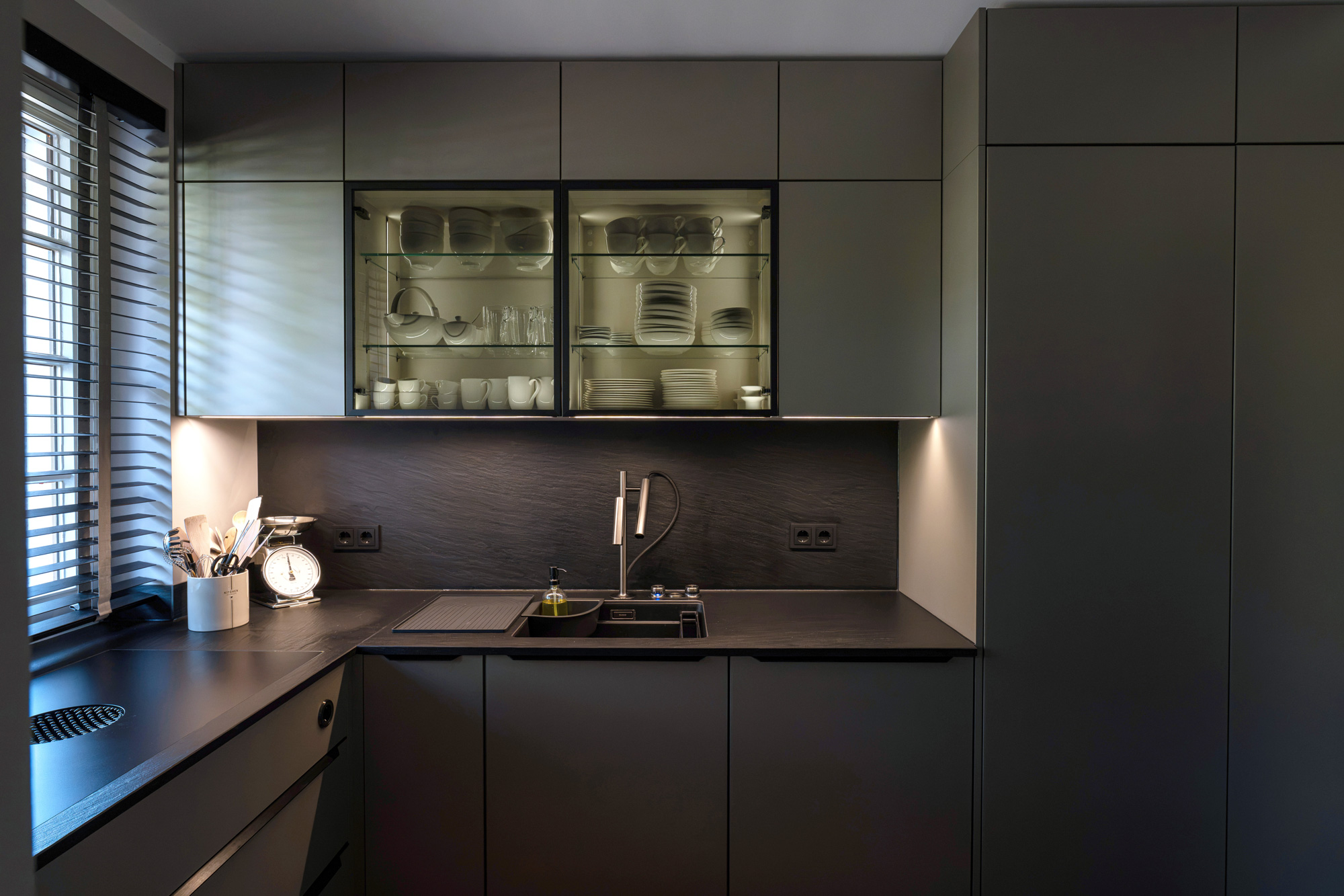 Moderne, dunkelgraue Küche mit beleuchteten Glasschränken, in denen weißes Geschirr steht, sowie einer dunklen Arbeitsplatte mit integriertem Spülbecken.
